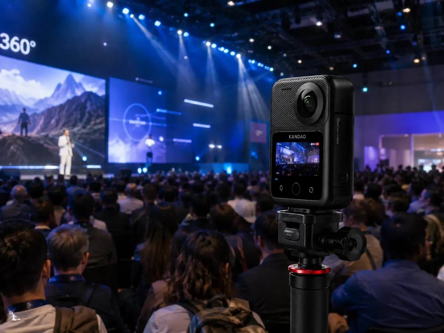 Caméra 360° Kandao sur trépied au premier plan, conférence grand public avec scène éclairée et écran géant en fond